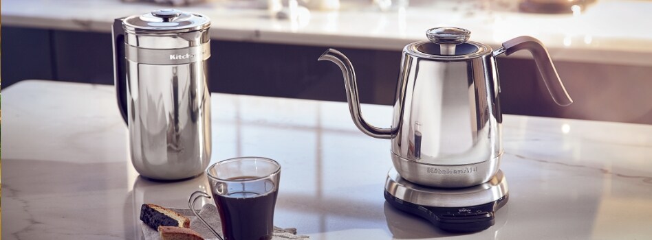 A cup of coffee on a dish cloth with two biscottis in front of a KitchenAid Precision Press Coffee Maker and a KitchenAid Gooseneck Kettle.